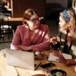 Two women seated at a table, collaborating on a laptop, engaged in discussion and sharing ideas.